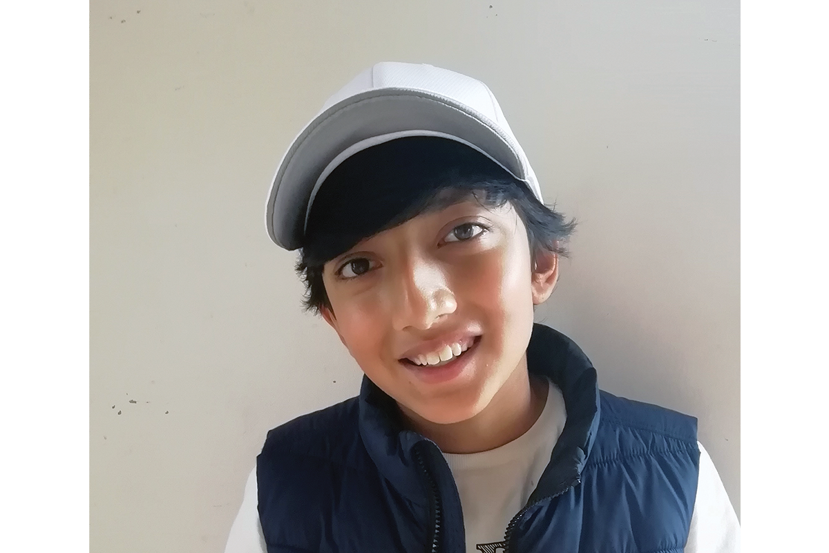 Head and shoulders shot of Aaryav, a young boy with a medium skin tone, wearing a white base ball cap and blue vest top. 